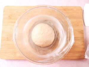 A ball of resting dough to make Three Kings bread sits in a greased glass bowl covered with plastic wrap, placed on a wooden cutting board.