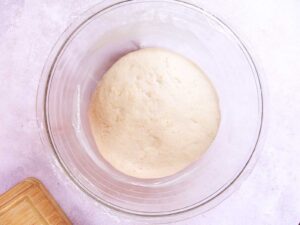 A ball of risen dough to make vegan rosca de reyes sits in the center of a clear glass bowl on a light-colored surface, with a corner of a wooden board visible in the bottom left.