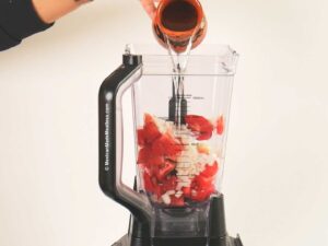 A hand pours water from a small cup into a blender filled with chopped tomatoes, garlic, and onions against a plain white background.