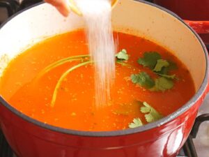 A hand pours salt into a red pot filled with sopa de estrellitas soup, garnished with fresh cilantro sprigs, on a stovetop.