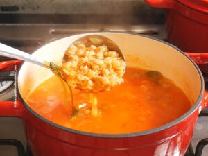 A ladle scoops up Mexican sopa de estrellas from a red pot. The soup, filled steams invitingly on the stove.