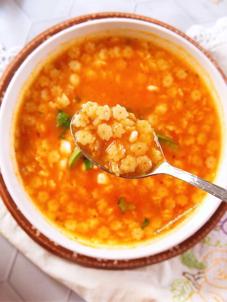 A close-up of a bowl of Mexican star pasta soup, a tomato-based dish with small star-shaped pasta, with a spoon lifting some sopita above the bowl. The soup is garnished with fresh herbs.