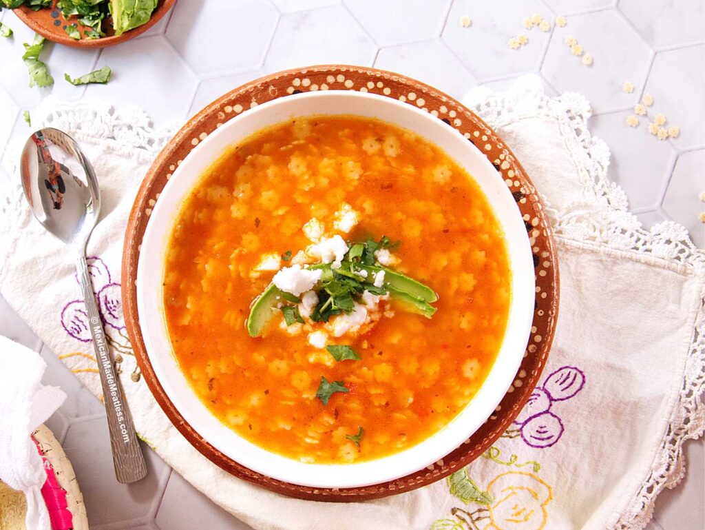 A bowl of orange-colored Mexican star soup with cheese crumbles, cilantro, and avocado slices on top, placed on a decorated plate and embroidered cloth, with a spoon and scattered ingredients nearby.