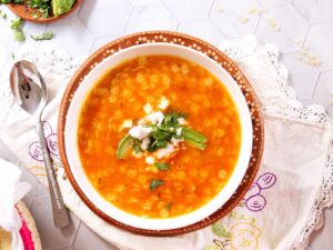 A bowl of orange-colored Mexican star soup with cheese crumbles, cilantro, and avocado slices on top, placed on a decorated plate and embroidered cloth, with a spoon and scattered ingredients nearby.