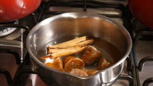 A stainless steel pot on a stove contains water, chunks of piloncillo (unrefined cane sugar), and cinnamon sticks, heating together. This is for making piloncillo syrup to drizzle over buñuelos.