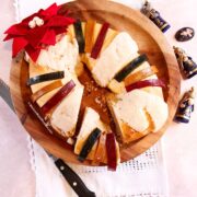 A sliced Three Kings Bread topped with colorful fruit strips sits on a wooden platter, decorated with a red poinsettia flower. A knife and small figurines are nearby on a light tablecloth.