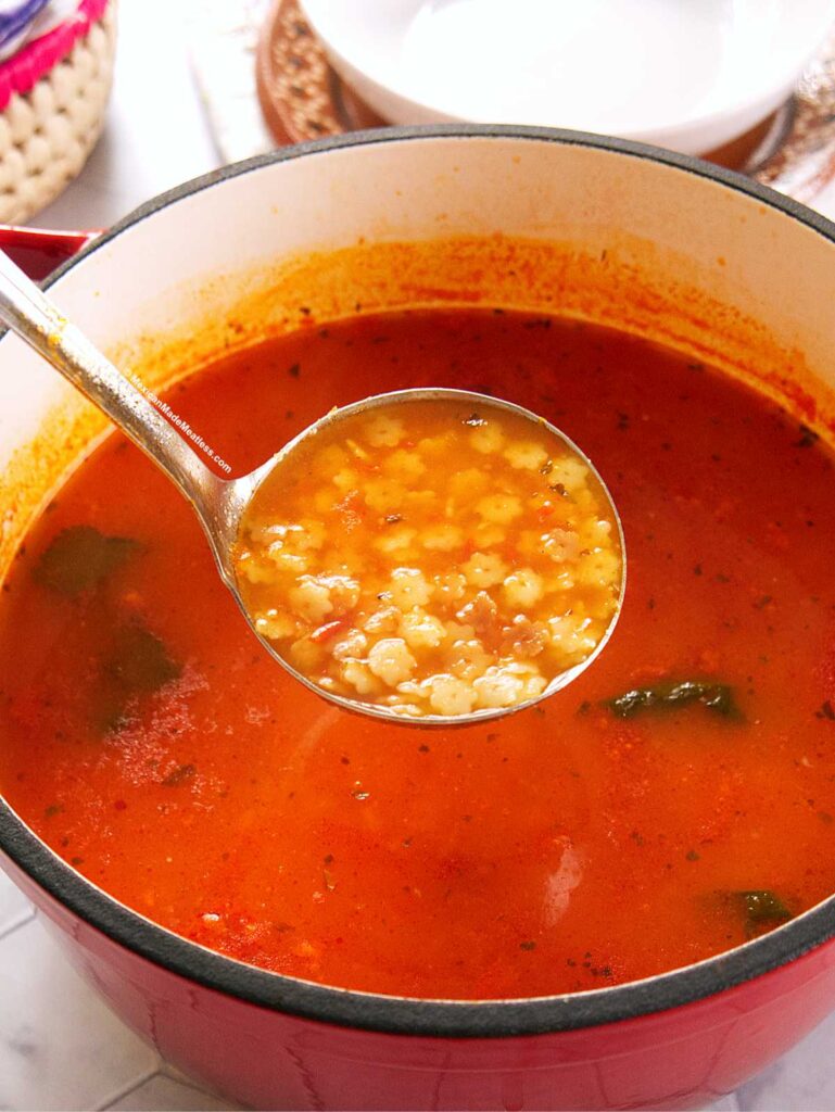 A metal ladle holds a serving of Mexican sopa de estrellitas soup—tomato-based with small star-shaped pasta and herbs—above a red pot filled with the same soup. White bowls are in the background.