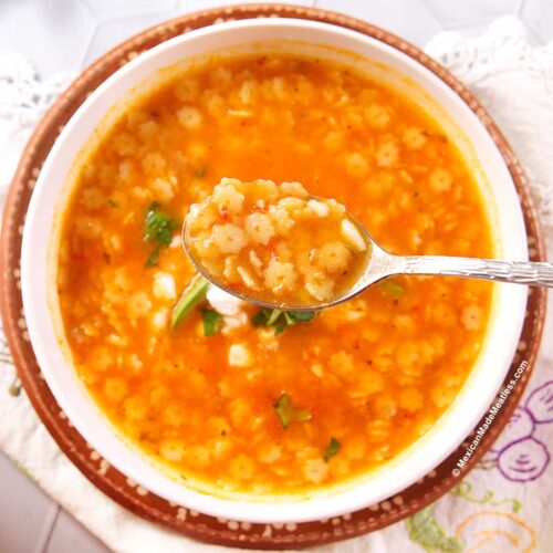 A bowl of Mexican sopa de estrellitas, or star soup, made with small star-shaped pasta, garnished with avocado, cheese, and cilantro. A spoon is held above the bowl, showing a portion of the Mexican tomato soup and pasta. The bowl sits on a brown plate on a patterned napkin.