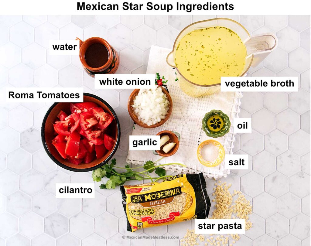 Labeled ingredients for Mexican star soup on a white surface: water, vegetable broth, oil, salt, garlic, diced white onion, chopped Roma tomatoes, cilantro, and a package of star-shaped pasta.