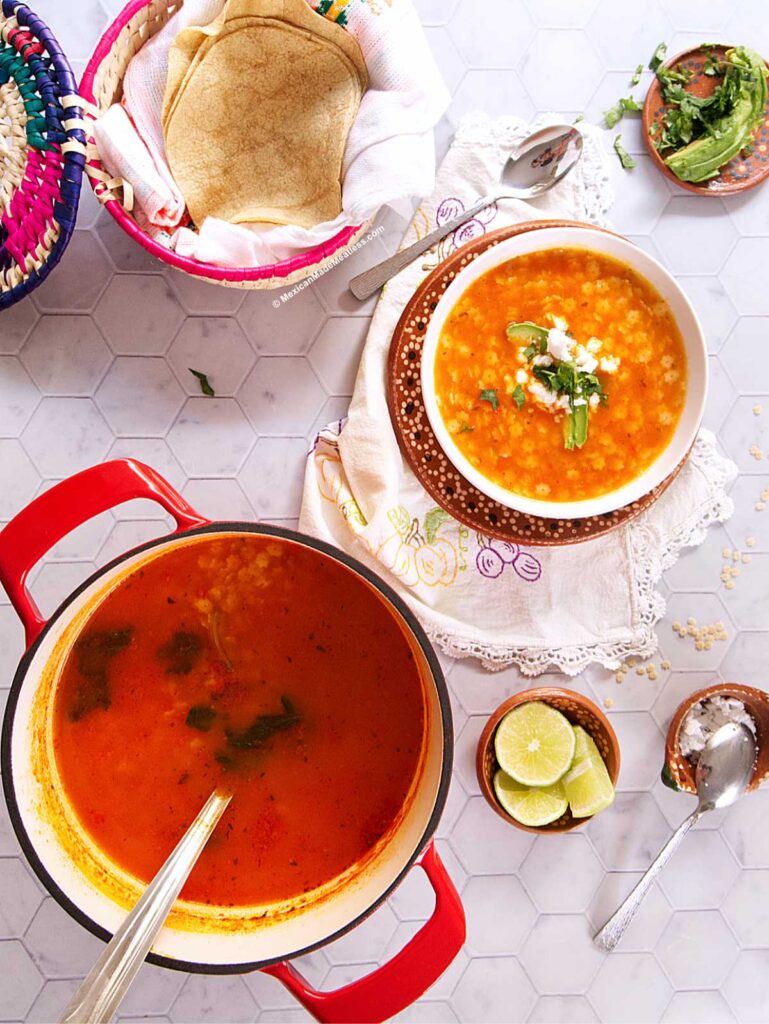 A red pot of soup sits on a hexagon-tiled surface next to a bowl of star soup topped with cilantro, lime wedges, a spoon, fresh herbs, and a basket of tortillas. The scene is bright and inviting.