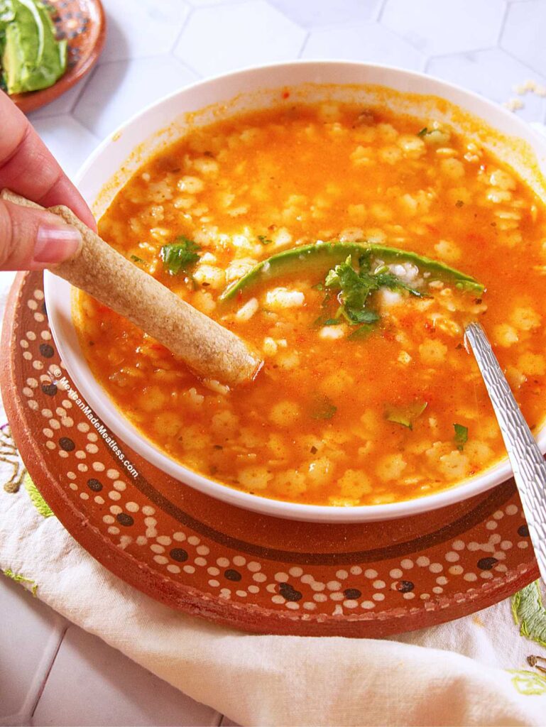 A bowl of tomato-based sopa de estrellas garnished with cilantro and a slice of avocado. A hand dips a rolled tortilla into the soup, and a spoon rests in the bowl, which sits atop a decorative plate.