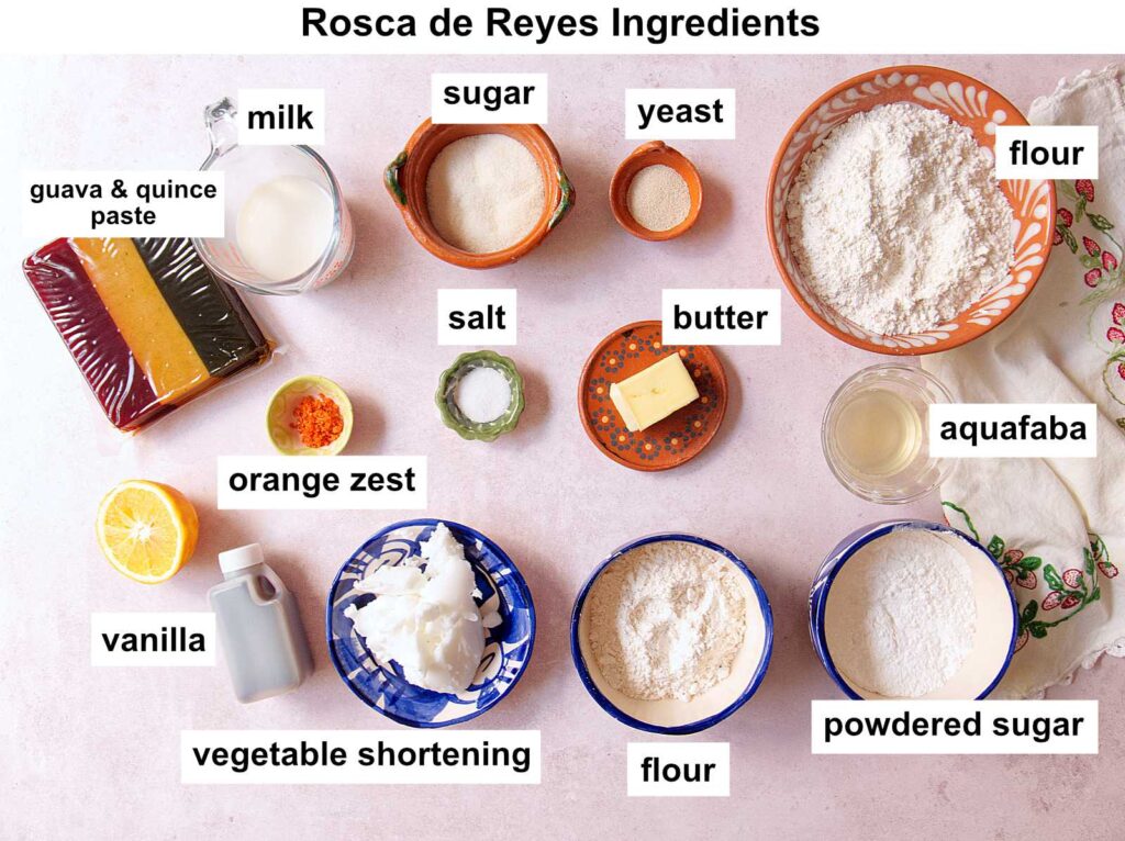 Rosca de Reyes ingredients arranged on a light surface, including flour, sugar, yeast, milk, butter, aquafaba, salt, orange zest, guava & quince paste, vanilla, powdered sugar, and vegetable shortening, each labeled.