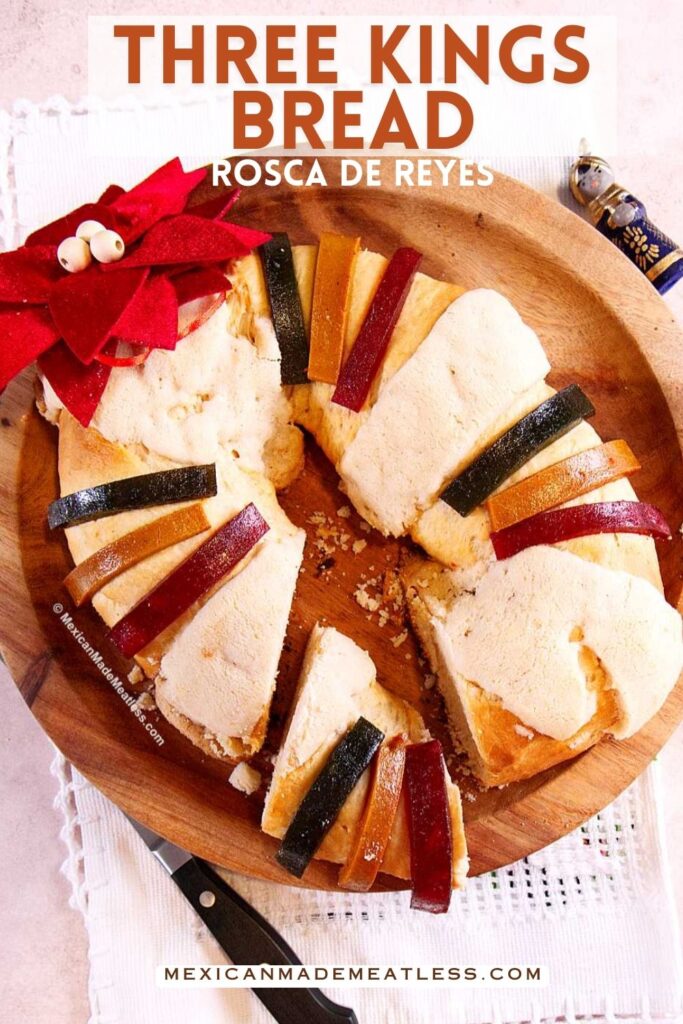A sliced round Rosca de Reyes bread topped with colorful candied fruit sits on a wooden platter. A red poinsettia and a small figurine are beside the bread. Text reads “Three Kings Bread Rosca de Reyes.”.