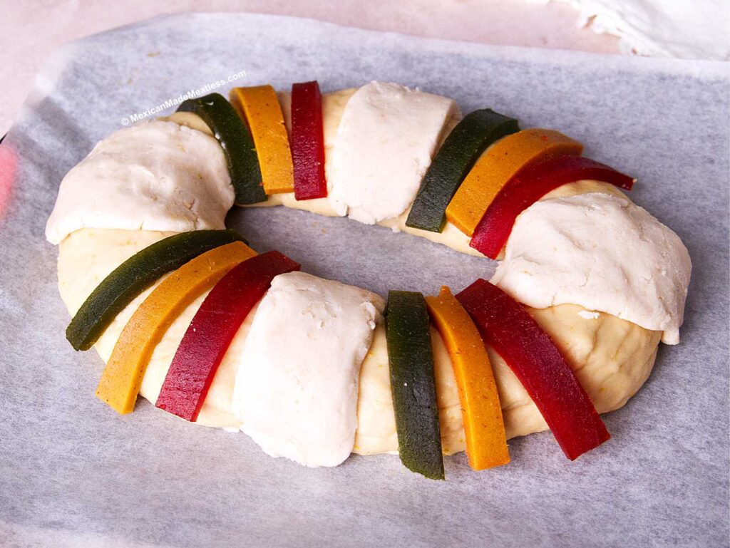 An oval-shaped Proofing Three Kings Bread dough ring on parchment paper, decorated with alternating strips of red, green, and yellow candied fruit paste, and topped with sections of sugar topping.
