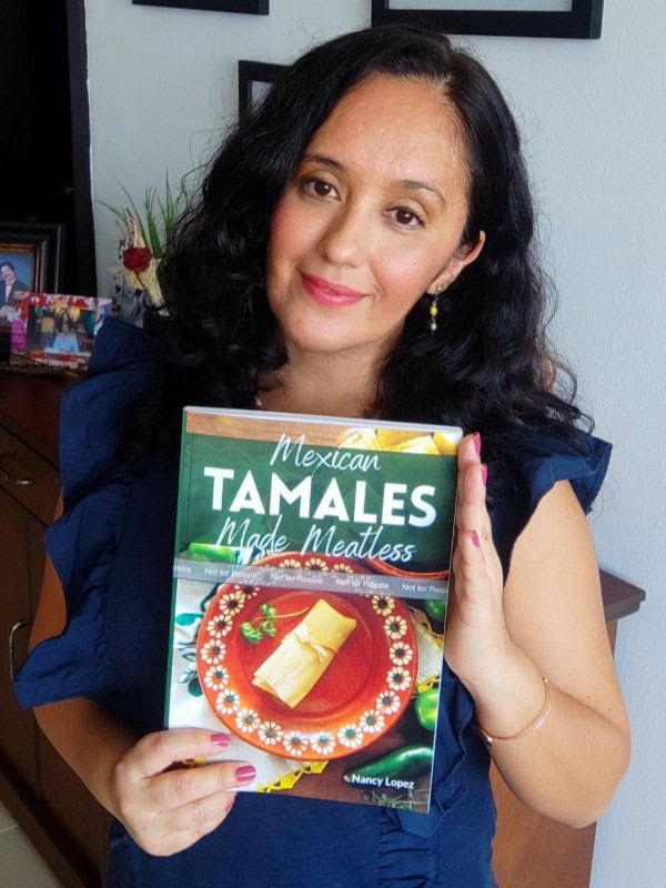Nancy Lopez, food blogger and cookbook author, stands indoors with a soft smile, holding up her cookbook "Mexican Tamales Made Meatless," featuring a tasty tamale on the cover. Her long dark hair frames her face warmly.