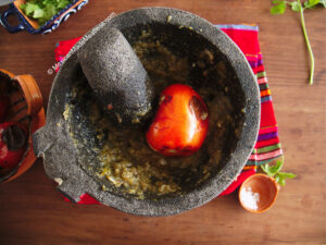A stone mortar with a pestle, roasted tomato, and green salsa on a colorful cloth, surrounded by herbs and a small bowl of salt on a wooden table.