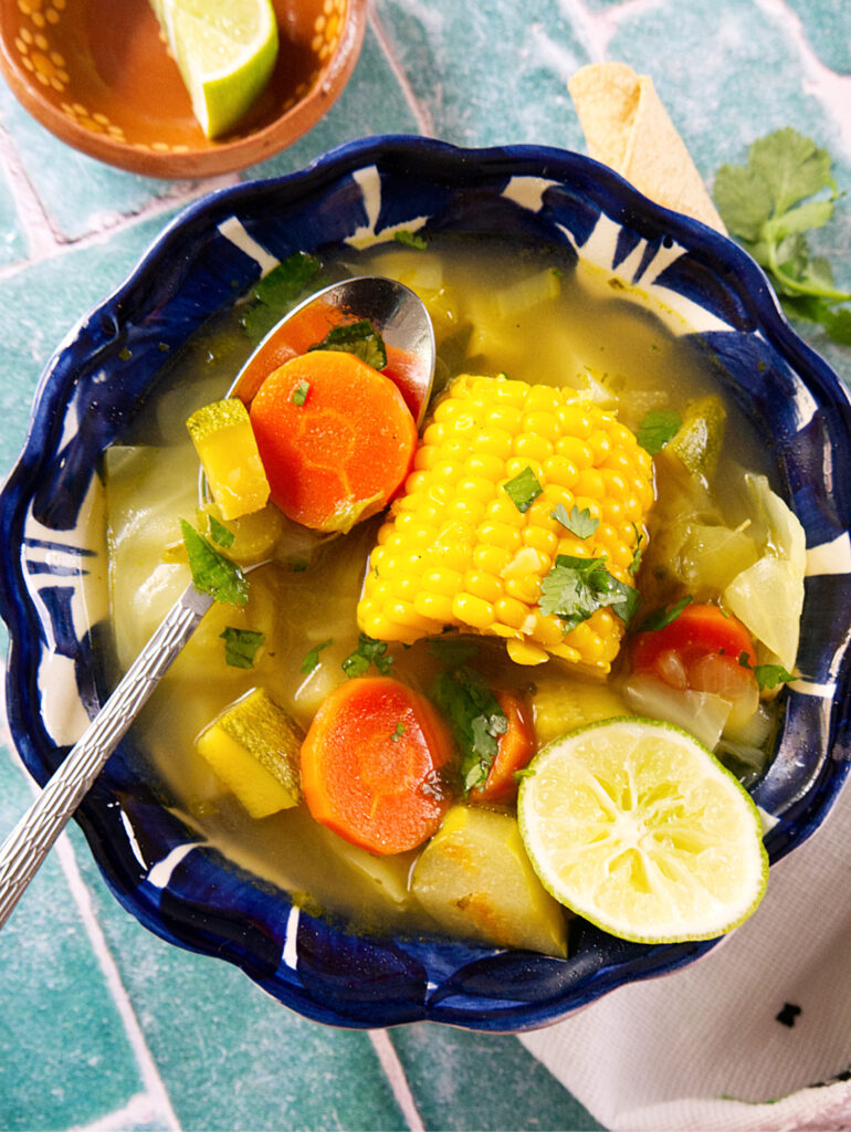 A bowl of Mexican vegetable soup with a corn cob, carrot slices, zucchini, and herbs, garnished with cilantro and a half lime, with a spoon resting inside the blue patterned bowl. A wedge of lime is in a dish nearby.