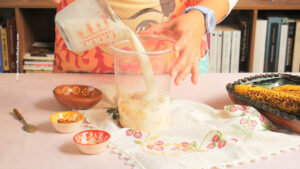 A person pours milk from a measuring cup into a blender jar with bananas inside, preparing a Mexican licuado de plátano. Various small bowls with spices, a spoon, and overripe bananas sit on a floral-embroidered cloth. Books are in the background.