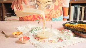 A person wearing a colorful apron pours a creamy Mexican banana smoothie from a pitcher into a glass on a table with a decorative cloth, two small bowls, and a platter of ripe bananas. Books are visible in the background.