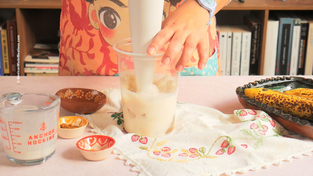 A person uses an immersion blender to mix ingredients for a Mexican banana smoothie in a tall container. On the table are milk, spices, ripe bananas, and decorative cloth, with books in the background.
