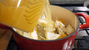 A close-up of vegetable broth being poured from a measuring container into a red pot on a stove, filled with cabbage, corn, and bay leaves.