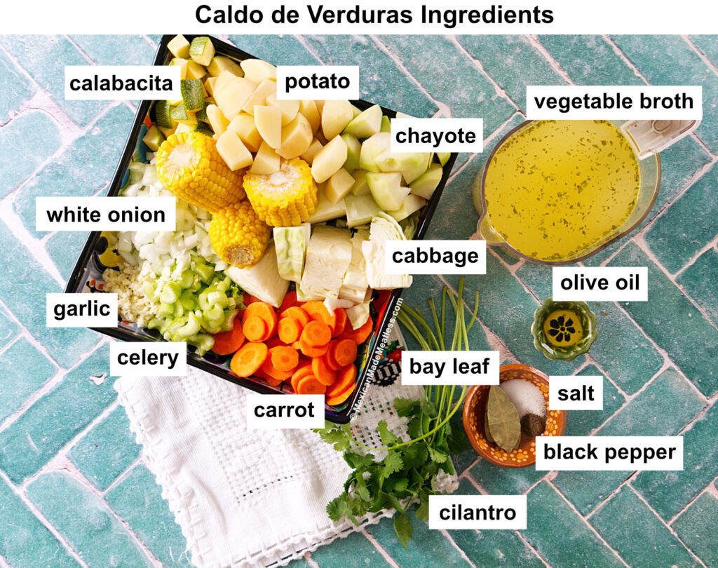 Labeled ingredients for caldo de verduras on a tiled surface, including chopped squash, potato, chayote, corn, onion, garlic, celery, carrot, cabbage, vegetable broth, olive oil, bay leaf, salt, black pepper, and cilantro.