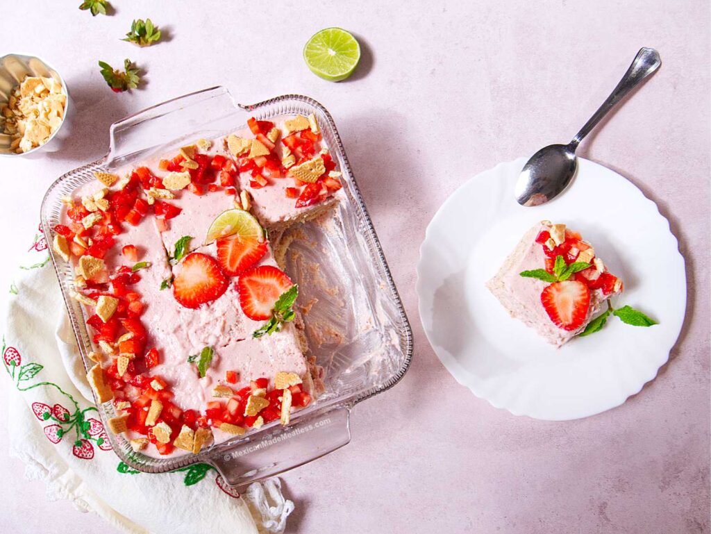 A glass baking dish holds a Mexican Strawberry Carlota, a no-bake icebox cake topped with chopped strawberries and mint. A slice is served on a white plate with a spoon, while lime and nuts rest nearby on the light pink background.