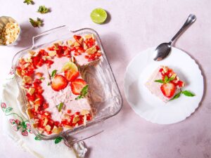 A glass baking dish holds a Mexican Strawberry Carlota, a no-bake icebox cake topped with chopped strawberries and mint. A slice is served on a white plate with a spoon, while lime and nuts rest nearby on the light pink background.