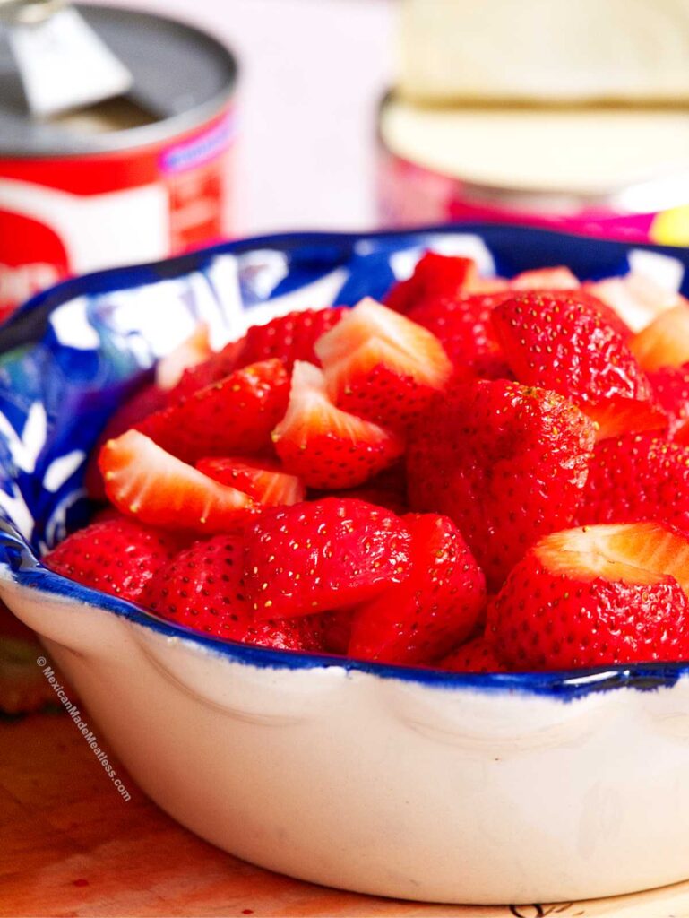 A bowl filled with sliced fresh strawberries sits on a table, with a blue and white rimmed edge. Blurred cans are visible in the background.