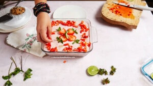 A hand places sliced strawberries on top of a Mexican Strawberry Carlota, a no-bake icebox cake, in a glass dish. Nearby are a cutting board with chopped strawberries, a lime, mint stems, crumbled cookies, and a white plate on a light surface.