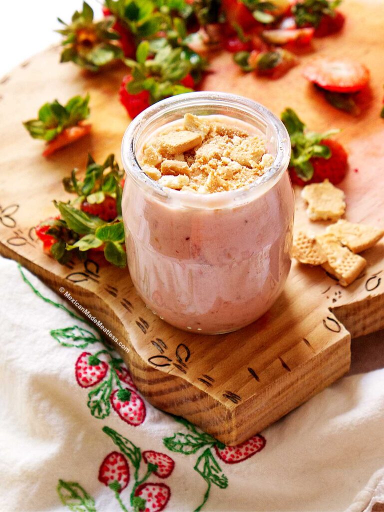 A glass jar of creamy Carlota de Fresa in jar for individual servings is topped with crushed cookies, sitting on a wooden board surrounded by fresh strawberries and leafy tops on a white cloth embroidered with red strawberries.