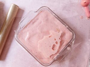 A glass square dish filled with light pink Mexican strawberry carlota sits on a light surface, and covered with plastic wrap ready to be refrigerated.