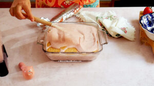 Nancy Lopez spreads a creamy pink mixture over layered cookies in a glass dish, preparing a classic Carlota de Fresa—an irresistible Mexican strawberry no-bake cake—amidst a cloth napkin, covered dish, and pink pig-shaped timer on the kitchen counter.