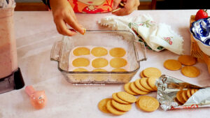 A person places round cookies in a glass baking dish lined with parchment paper, preparing a Mexican strawberry icebox cake. More cookies, a cloth napkin, and a blender with a pink mixture are on the countertop nearby.