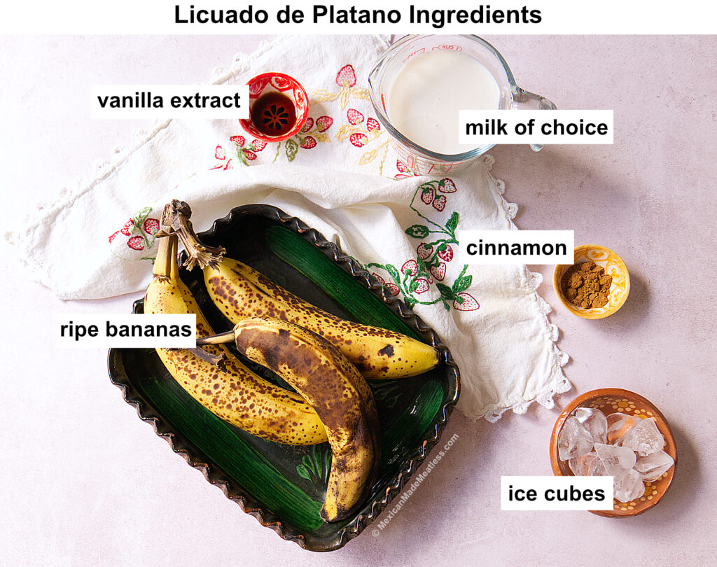 Overhead view of ingredients for licuado de plátano: ripe bananas in a green dish, vanilla extract in a small red bowl, milk in a glass measuring cup, ground cinnamon in a small bowl, and ice cubes in a dish.