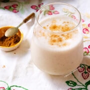 A glass of creamy licuado de platano, is topped with cinnamon and sits on a floral embroidered cloth next to a small yellow bowl filled with ground cinnamon and a spoon.