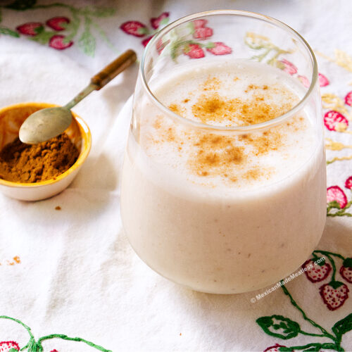 A glass of creamy licuado de platano, is topped with cinnamon and sits on a floral embroidered cloth next to a small yellow bowl filled with ground cinnamon and a spoon.