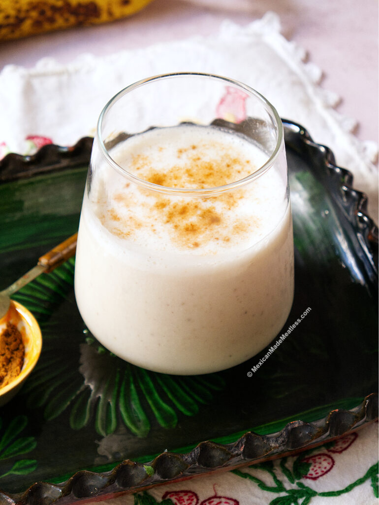A glass of creamy Mexican banana smoothie topped with a sprinkle of cinnamon sits on a green decorative tray, next to a small spoonful of ground canela. A white napkin is partially visible in the background.