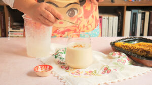 Nancy Lopez sprinkles ground cinnamon over Mexican banana smoothie. Ripe bananas and a small bowl are nearby, with books and shelves in the background.