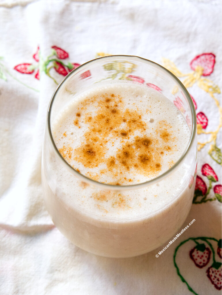 A creamy, frothy Mexican banana smoothie, or licuado de plátano, is served in a round glass on a white cloth decorated with embroidered strawberries and flowers, then topped with ground cinnamon.
