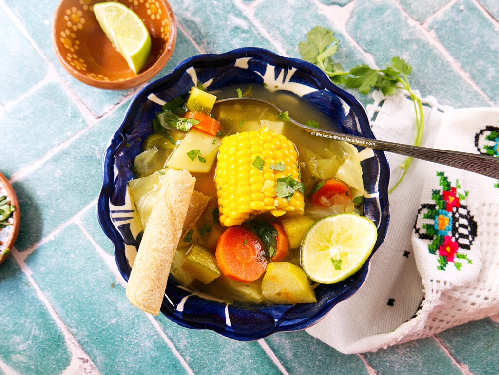 A bowl of caldo de verduras featuring corn on the cob, carrots, zucchini, and potatoes, garnished with cilantro and served with a lime wedge and a piece of bread on a blue patterned table.