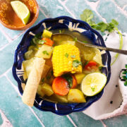 A bowl of caldo de verduras featuring corn on the cob, carrots, zucchini, and potatoes, garnished with cilantro and served with a lime wedge and a piece of bread on a blue patterned table.