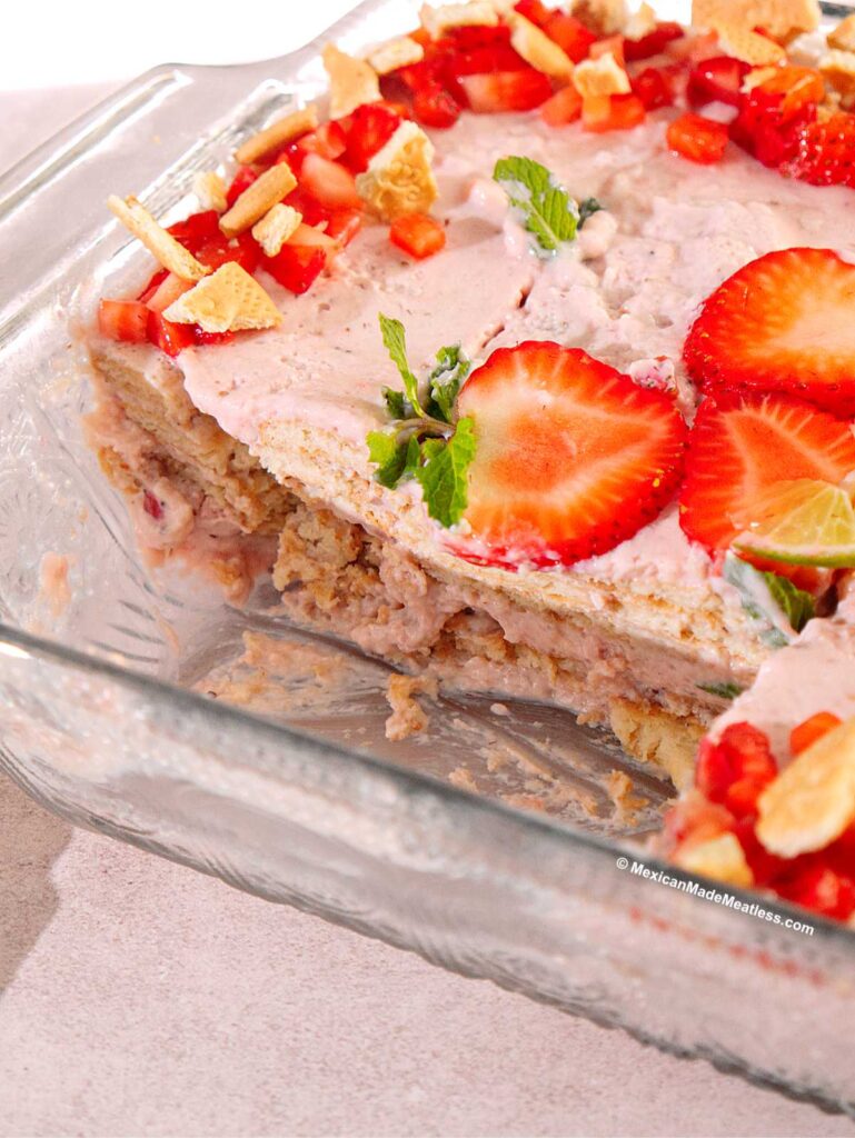 A glass baking dish with a partially served Carlota de fresa Mexican strawberry dessert made of cream, Maria cookies, and strawberries. Sliced strawberries, mint leaves, and cookie pieces decorate the top.
