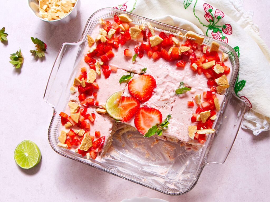 A glass baking dish with a creamy pink Strawberry Carlota dessert topped with chopped strawberries and Maria cookie pieces. Two halved strawberries and mint leaves are on top. A portion has been removed, showing a crumbly crust. Lime and strawberries nearby.