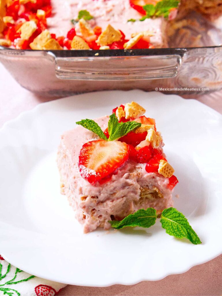 A slice of Carlota de fresa Mexican strawberry icebox cake made with Maria cookies and fresh strawberries sits on a white plate, topped with crumbled cookies and mint leaves, with a glass dish of the same dessert in the background.