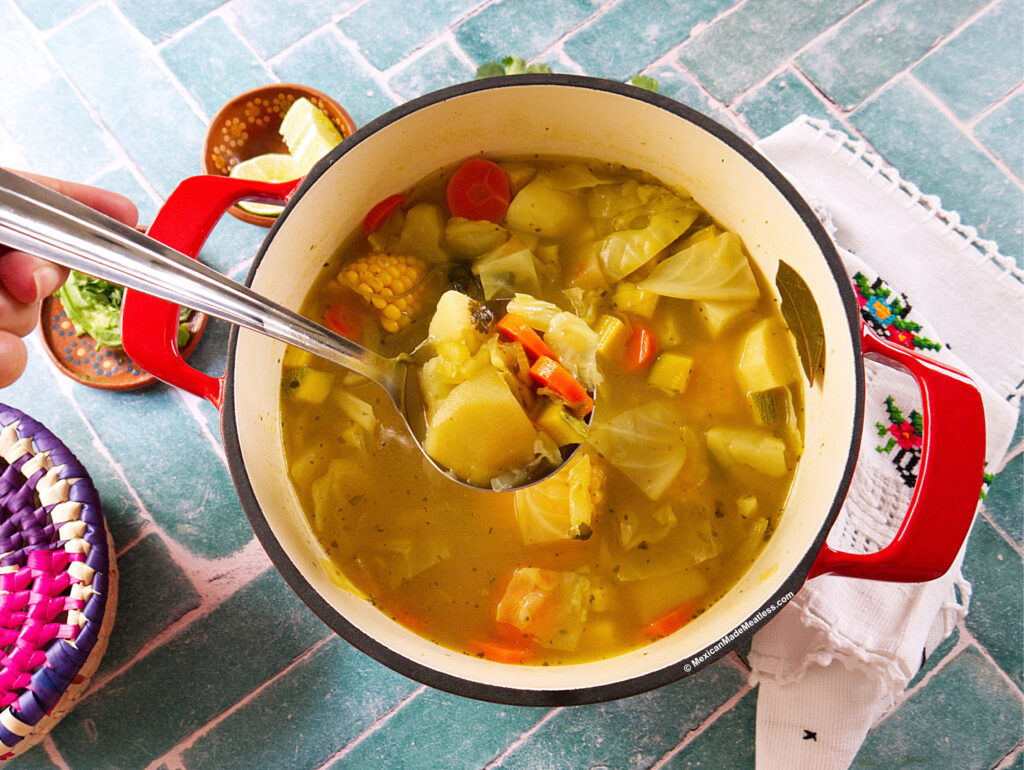 A hand holds a ladle scooping Mexican vegetable stew with potatoes, carrots, corn, and cabbage from a red pot on a blue tile surface, with a colorful woven basket and embroidered napkin nearby.