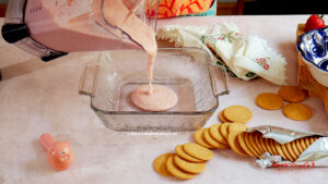 Nancy Lopez pours a pink carlota de fresa cream mixture from a blender into a glass baking dish. Surrounding the dish are stacked Maria cookies, an opened package, a pig-shaped spoon, and a floral napkin on a light surface.