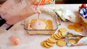 A blender cup pours pink cream over a layer of round cookies in a glass dish. More cookies and a foil wrapper are nearby on a light countertop, with colorful napkins and a pig-shaped timer in the scene.