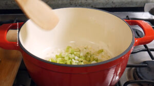 A red pot on a stove with chopped onions and celery being stirred with a wooden spatula. The pot is placed on a burner, and the vegetables are beginning to cook.