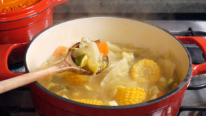 A red pot on a stovetop contains simmering caldo de verduras with corn, carrots, zucchini, and cabbage. A wooden ladle lifts some of the soup out of the pot as steam rises from the hot broth.